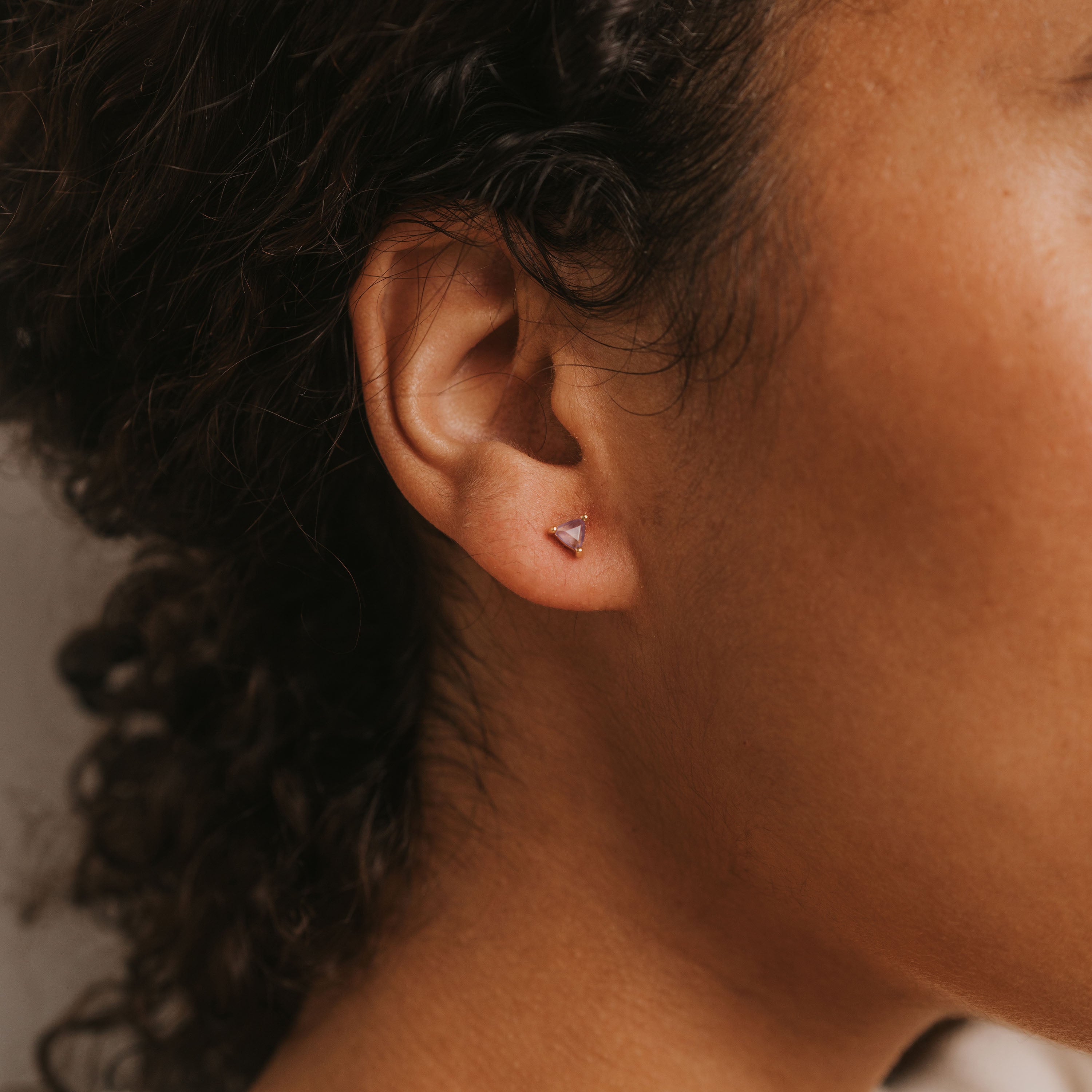 Close-up of a person’s ear wearing an Amethyst Mini Energy Gems - Healing stud earring, featuring a small triangular purple gem on sterling silver posts. The person has curly brown hair and warm-toned skin.