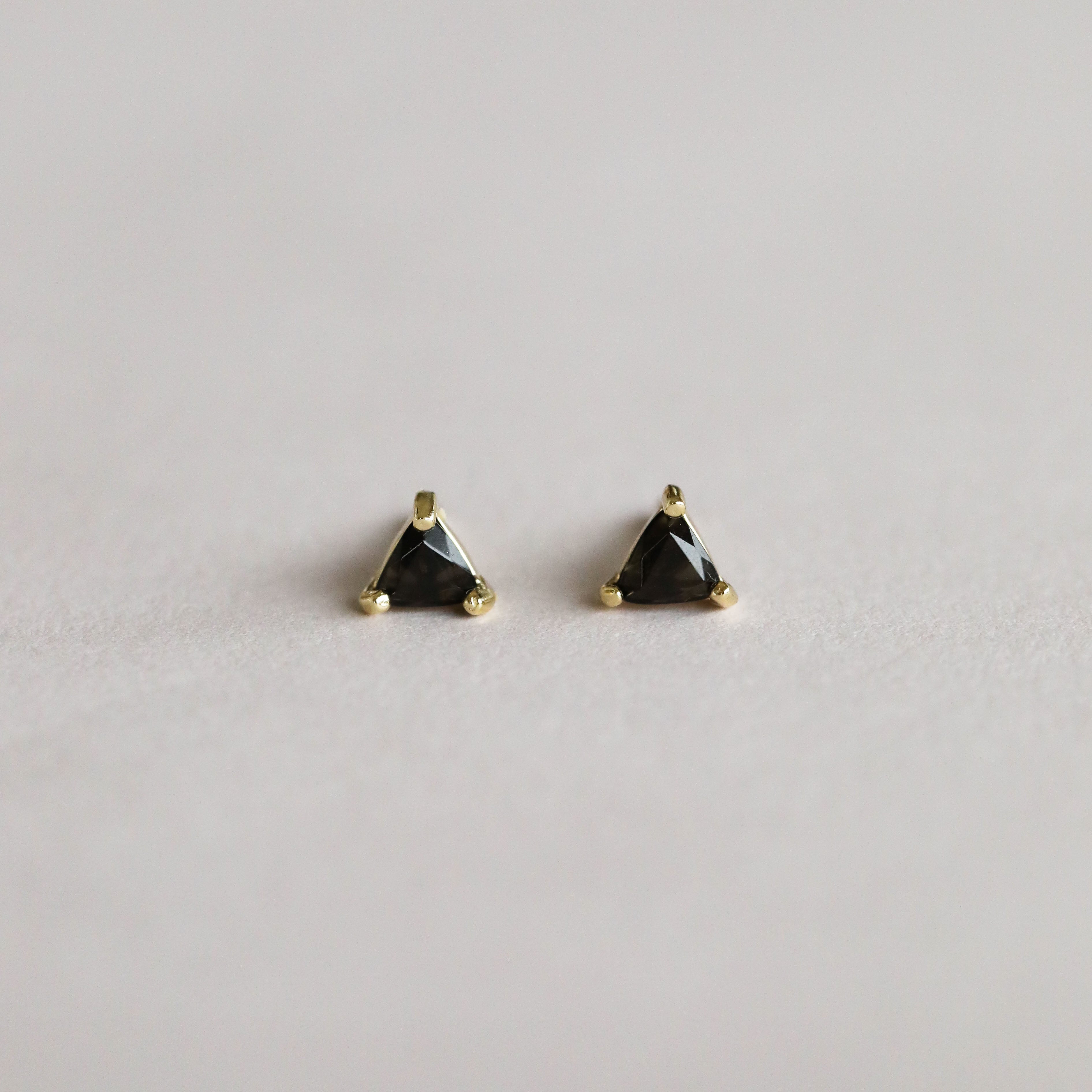 A pair of Black Tourmaline Mini Energy Gems - Strength studs, small and triangular with sterling silver posts and 18k gold plating, displayed on a plain light gray background.