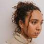 A woman with curly hair in a bun and a cream turtleneck looks right, showcasing elegant Black Tourmaline Mini Energy Gems - Strength studs with sterling silver posts against a plain white background.