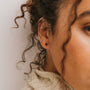 Close-up of a woman's ear wearing the Black Tourmaline Mini Energy Gems - Strength earring, featuring 18k gold plating. Her curly brown hair and cream-colored sweater create a warm, softly blurred background.