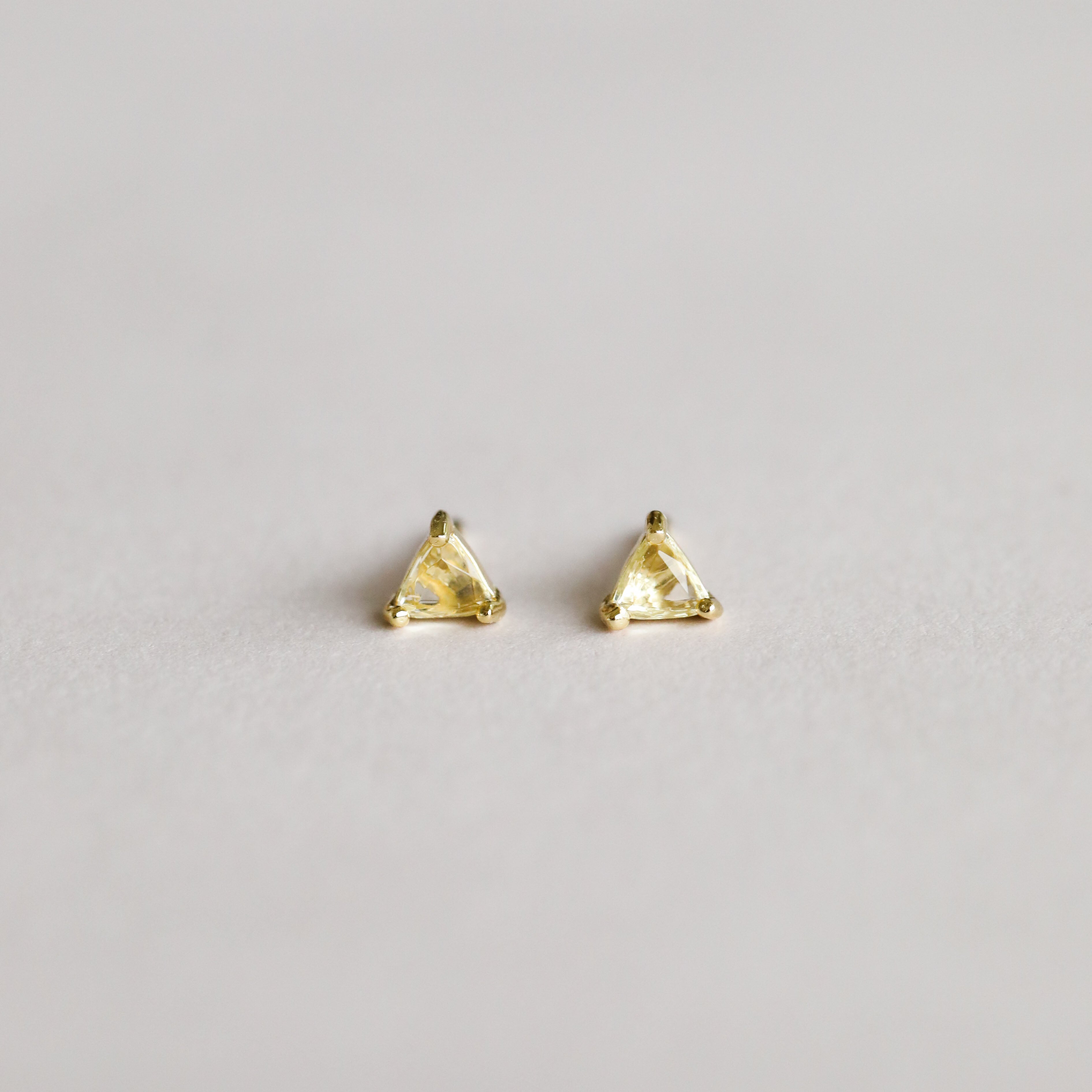 A pair of Citrine - Mini Energy Gems - Success studs, featuring small triangular yellow gemstones with 18k gold plating and sterling silver posts, shown on a plain light gray background.