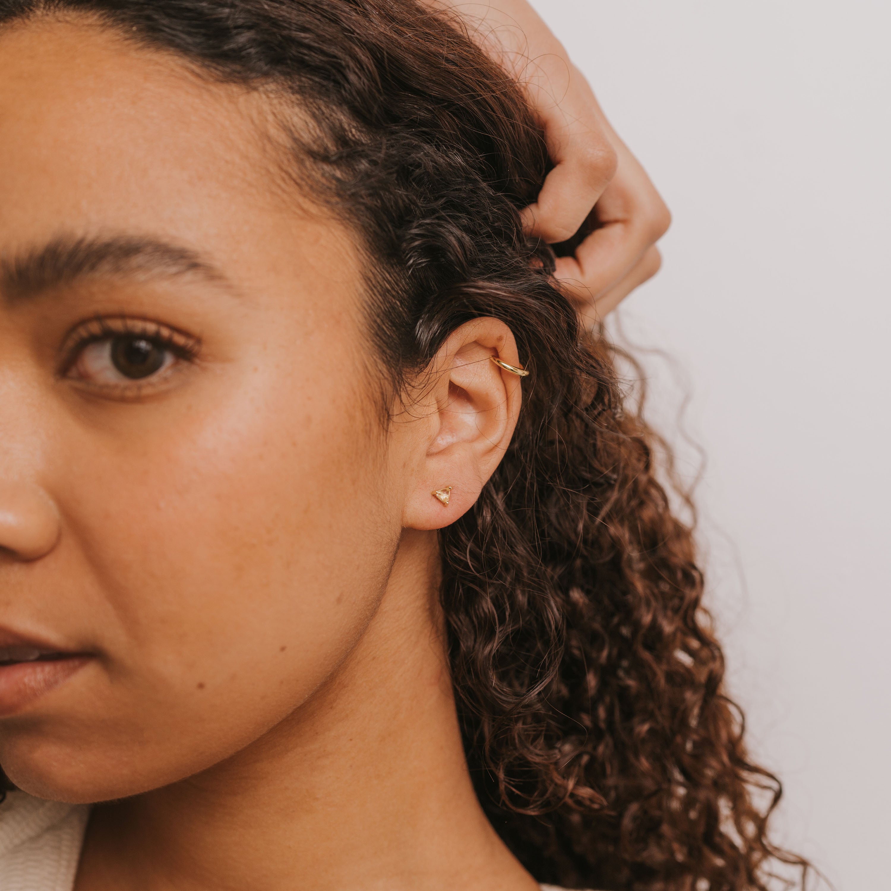 A woman with curly hair wears the Citrine - Mini Energy Gems - Success earrings, featuring 18k gold plating and sterling silver posts, with her hand touching her hair near her ear. The background is plain white.