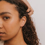 A woman with curly hair wears the Citrine - Mini Energy Gems - Success earrings, featuring 18k gold plating and sterling silver posts, with her hand touching her hair near her ear. The background is plain white.