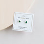 A pair of Green Onyx Mini Energy Gems - Abundance studs, featuring triangular green onyx set in 18k gold plating, are displayed on a white card with product details against a light neutral background.