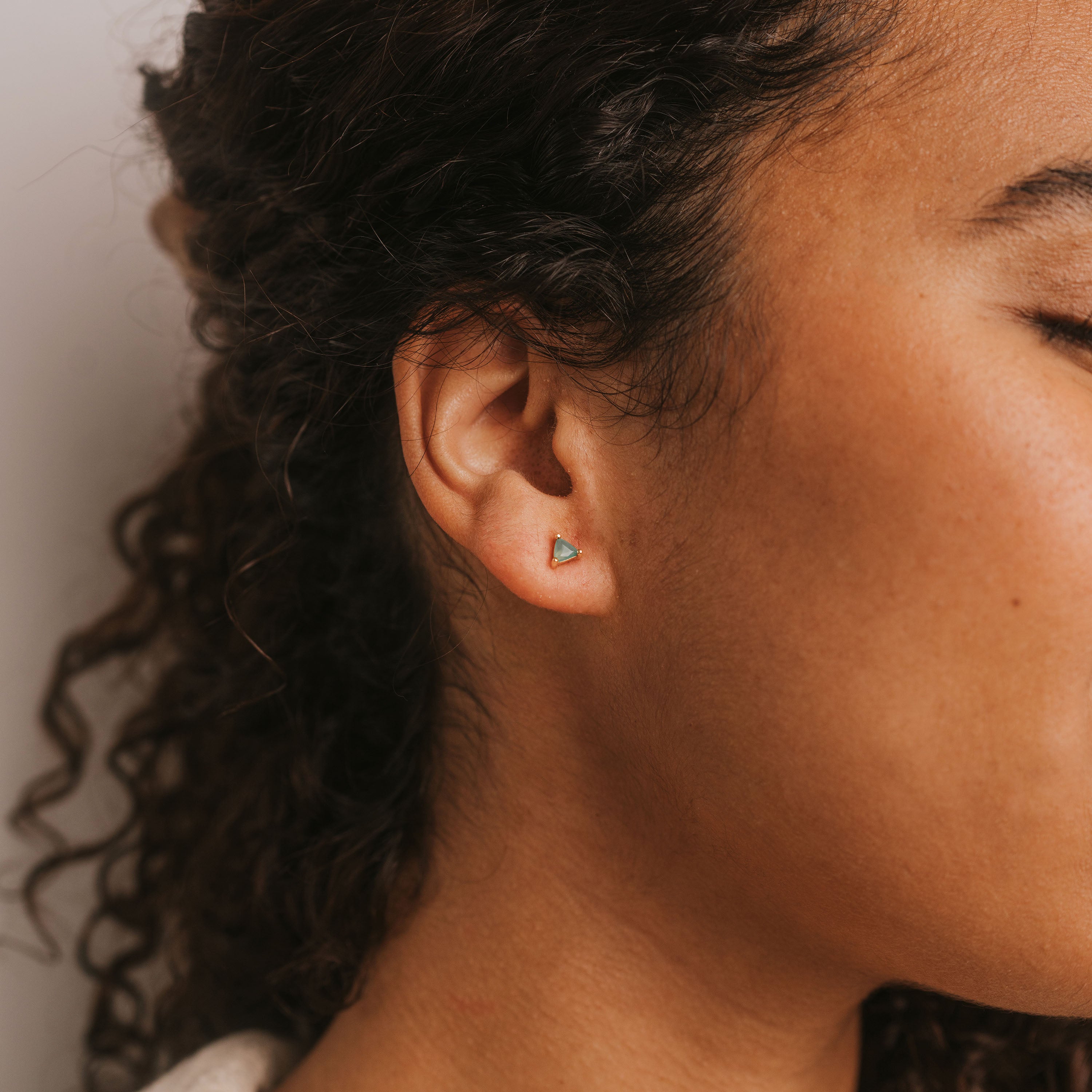 Close-up of a person's ear with Green Onyx Mini Energy Gems - Abundance studs. Their curly hair frames their face, eyes closed and skin smooth, expressing peace.