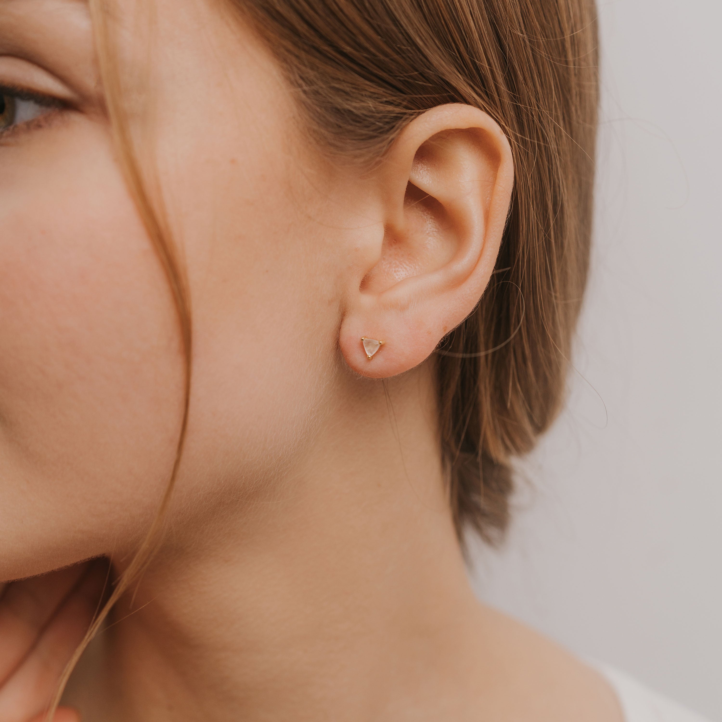 Close-up of a woman's ear wearing Rose Quartz Mini Energy Gems - Love studs with 18k gold plating and sterling silver posts. Part of her face, brown hair, and hand appear against a neutral background.