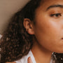Close-up of a person with curly hair wearing the Minimalist Bar, a small 18k gold plated hoop earring, in their left ear. The neutral background highlights their ear and cheek, drawing attention to the elegant gold accent.