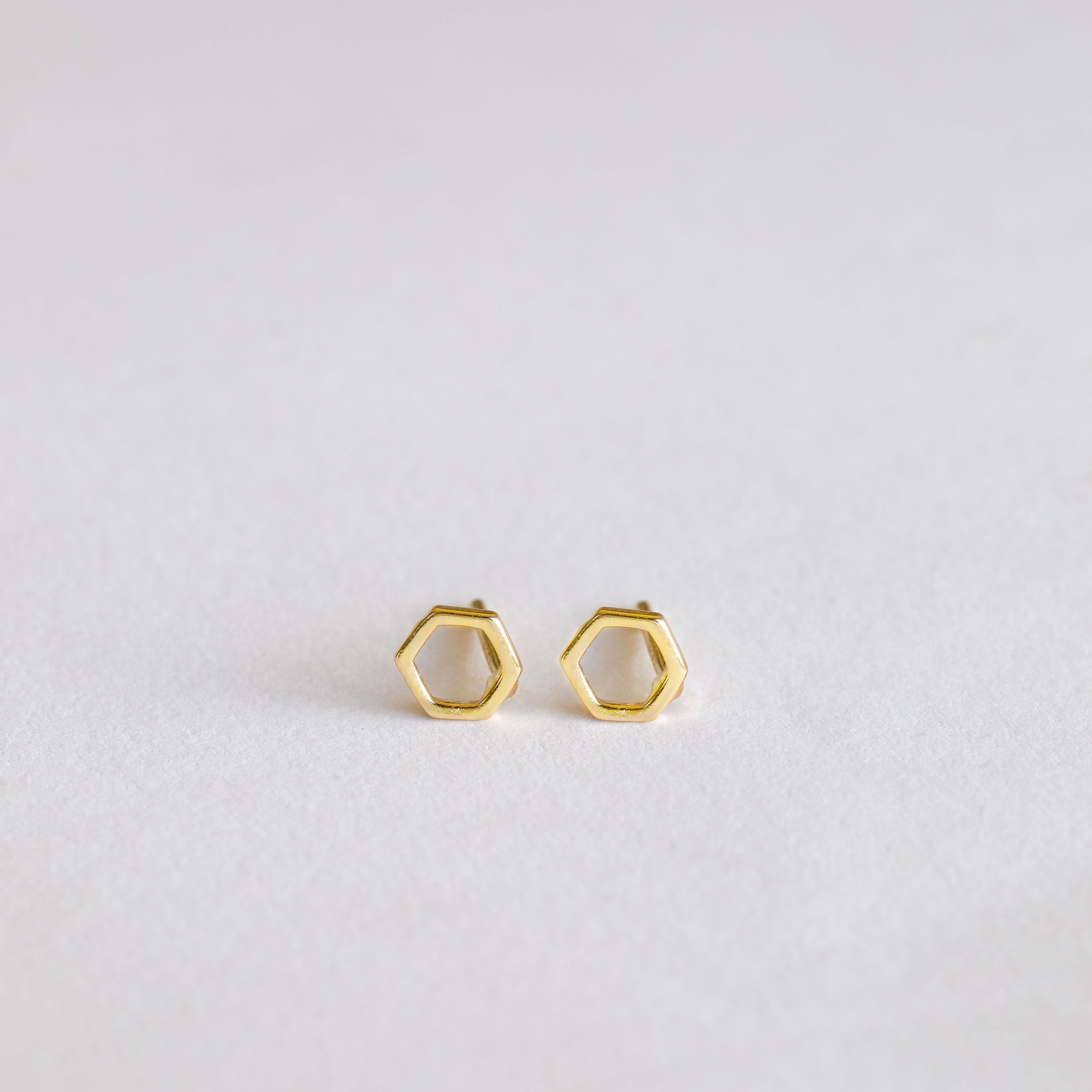 The Minimalist Hexagon earrings are a pair of small, gold plated hexagons shown on a plain, light background.