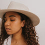 A woman with curly brown hair wears the Minimalist Hexagon sterling silver earrings, a cream wide-brimmed hat, and a white shirt. She gazes downward and to the side against a simple, light background.