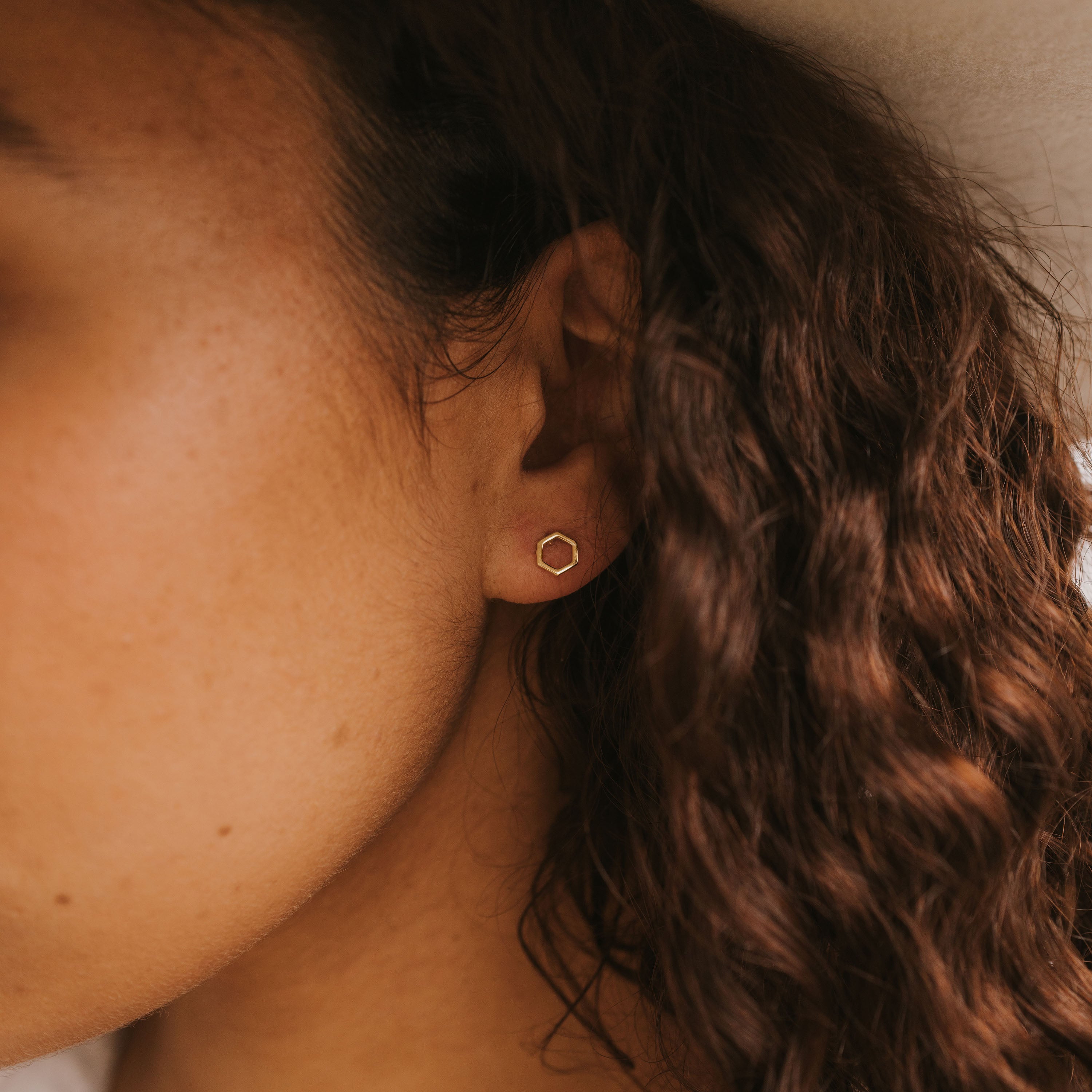 A close-up shows a person with curly brown hair wearing the Minimalist Hexagon, a small gold-plated earring. The background and part of their face are softly blurred.