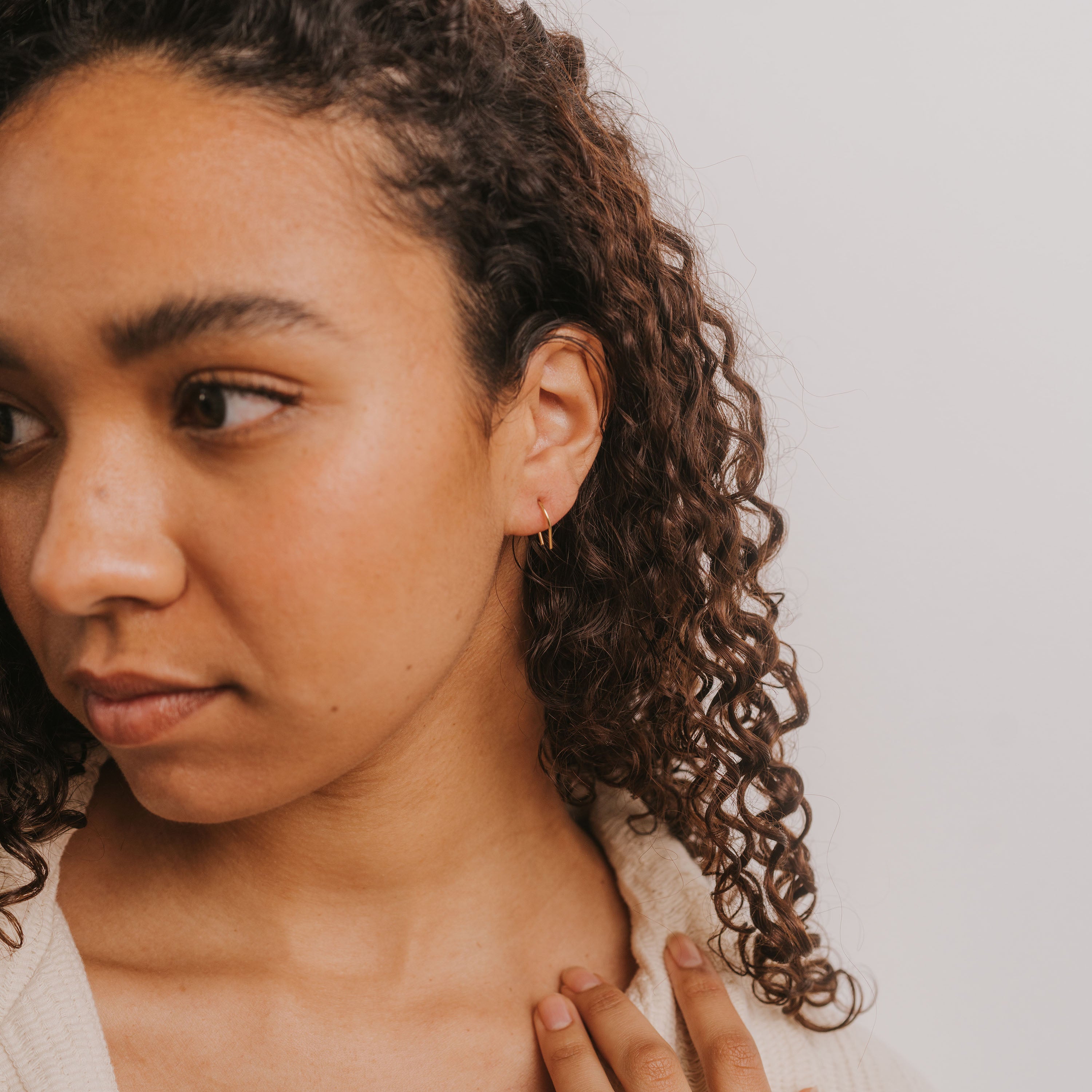 A woman with curly hair and medium skin tone looks to the side, wearing a cream top and Minimalist Horseshoe 18k gold plated earrings. Her hand rests near her collarbone against a plain background.