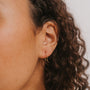 Close-up of a person’s ear with curly brown hair, wearing the Minimalist Horseshoe 18k gold plated hoop earring. A neutral, softly blurred background highlights the elegant jewelry detail.