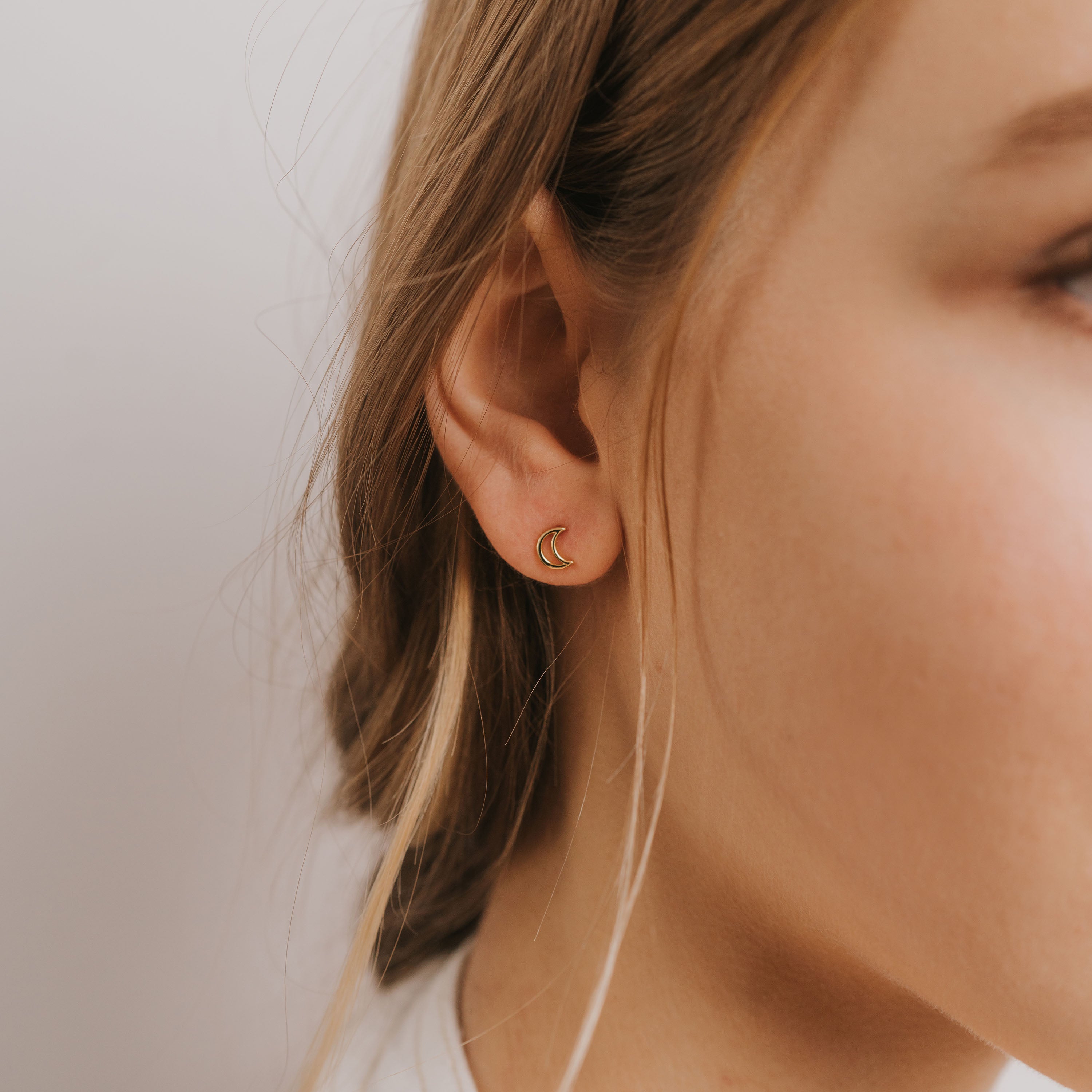 Close-up of a woman's ear wearing the Minimalist Moon—a small, 18k gold plated hollow moon earring. Her light brown hair is tucked behind her ear against a plain white background, emphasizing this elegant sterling silver jewelry.