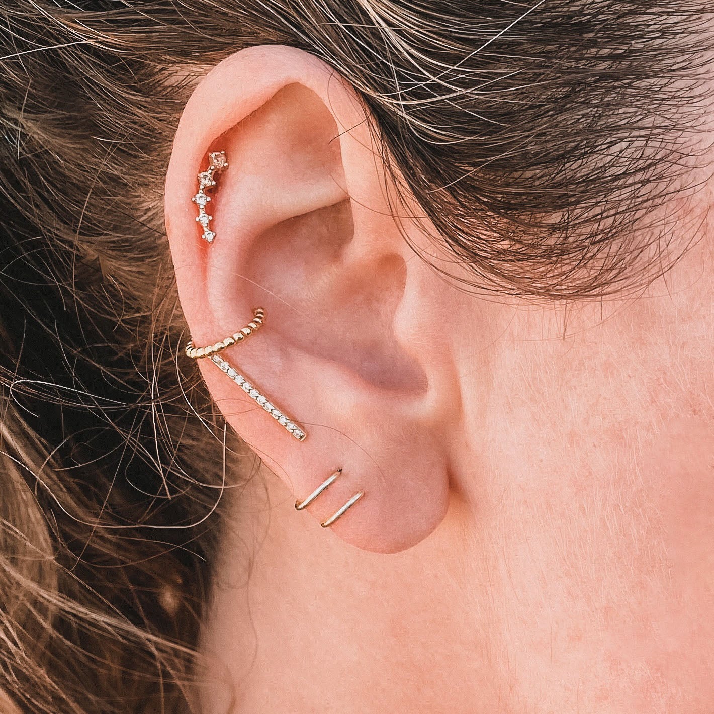 Close-up of an ear with multiple piercings wearing gold and diamond earrings, highlighted by the Minimalist Spiral—a spiral earring with 18k gold plating—adorned along the lobe, helix, and cartilage. Brown hair gently frames the ear.