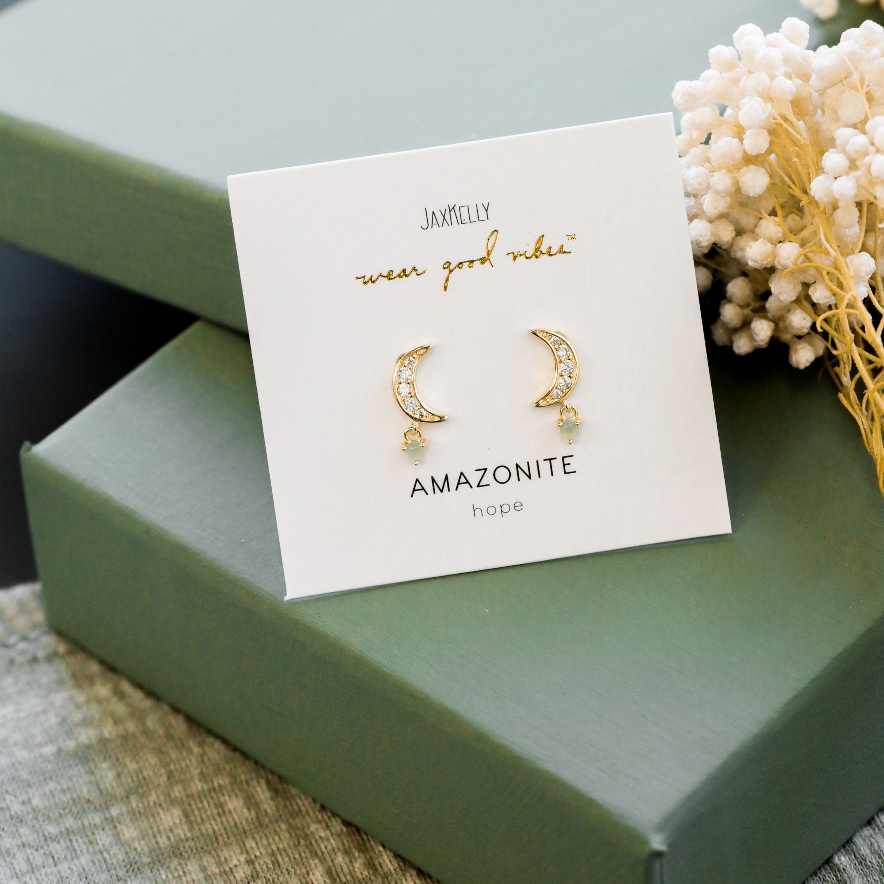 The Amazonite Moon Drop earrings in gold plated sterling silver feature small amazonite stones, displayed on a JaxKelly "wear good vibes" card that says "Amazonite hope," set atop a green gift box with white dried flowers.