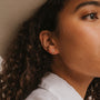 Close-up of a person with curly dark hair wearing the Amazonite Moon Drop, a small gold plated sterling silver moon stud earring. The focus is on the ear and earring, against a neutral background with part of a white shirt visible.
