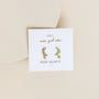 A white card displaying the Rose Quartz Moon Drop earrings—moon studs with small crystals and rose quartz stones, crafted from gold-plated sterling silver—set on a beige background. The card reads: JaxKelly, wear good vibes, ROSE QUARTZ, love.