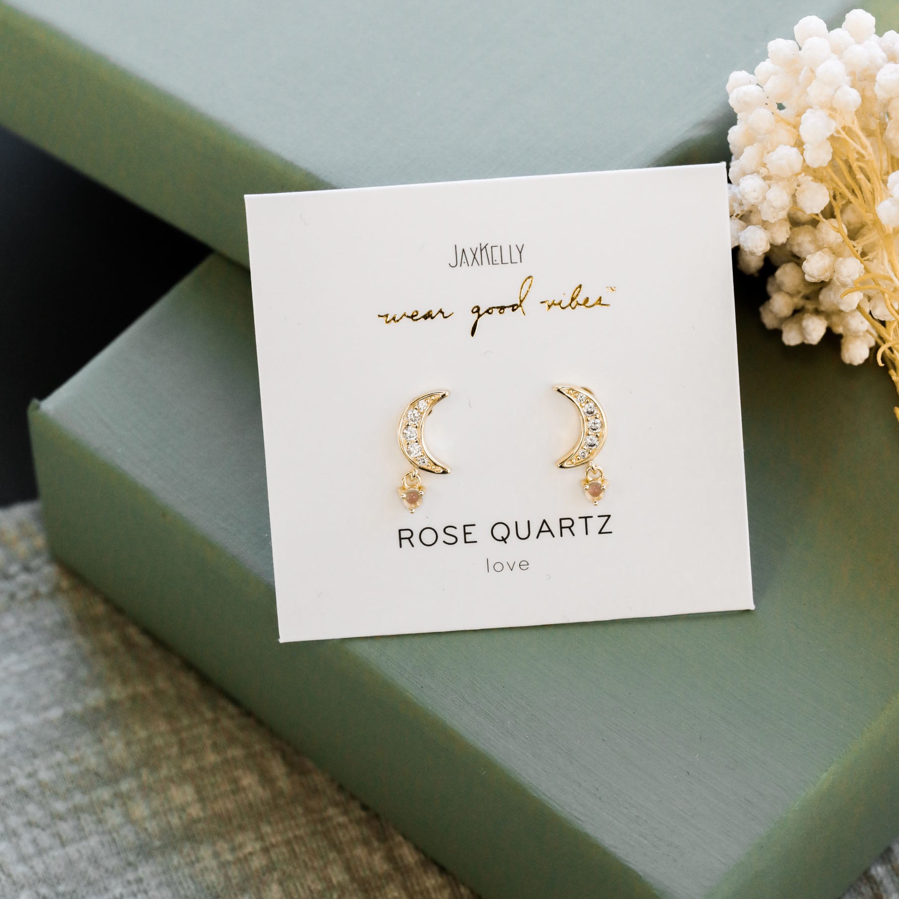 A card labeled JaxKelly displays the Rose Quartz Moon Drop: gold-plated sterling silver moon stud earrings featuring small gemstones and dangling rose quartz charms, set on green boxes beside white dried flowers.