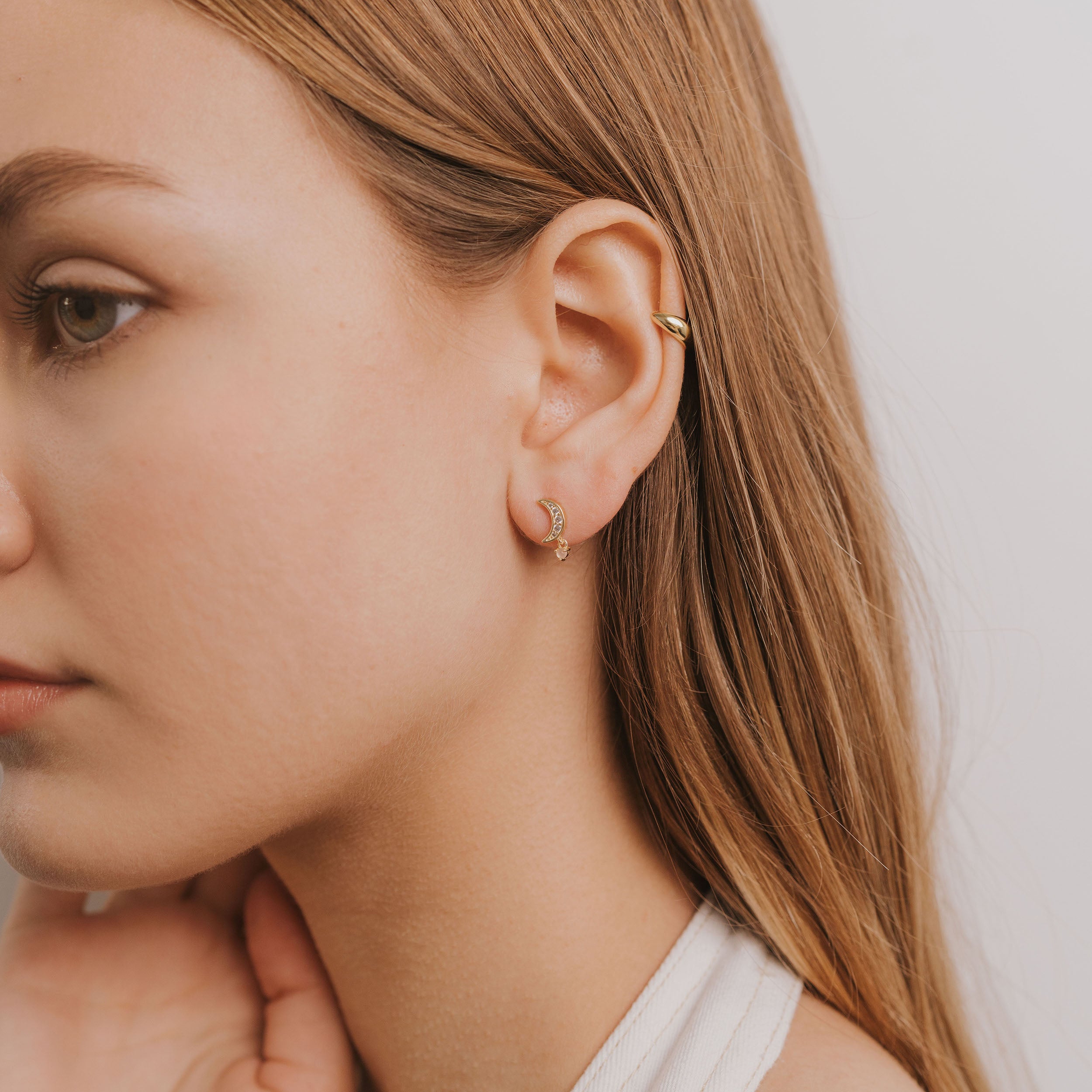 A young woman with long, straight light brown hair wears a small gold hoop earring, a gold ear cuff, and the delicate Rose Quartz Moon Drop jewelry. She faces slightly left, gently touching her neck against a plain white background.