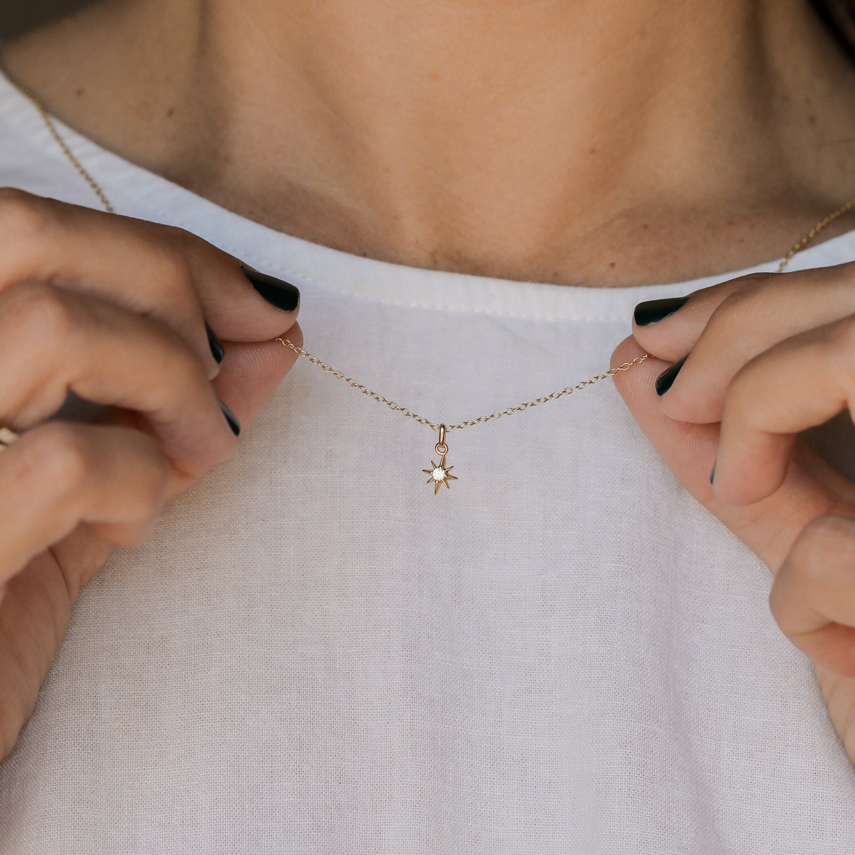 A person in a white top holds the Tiny Star Necklace—an elegant 18k gold plated piece with a small star-shaped CZ stone pendant—close to their neck, showing off their dark green painted nails.