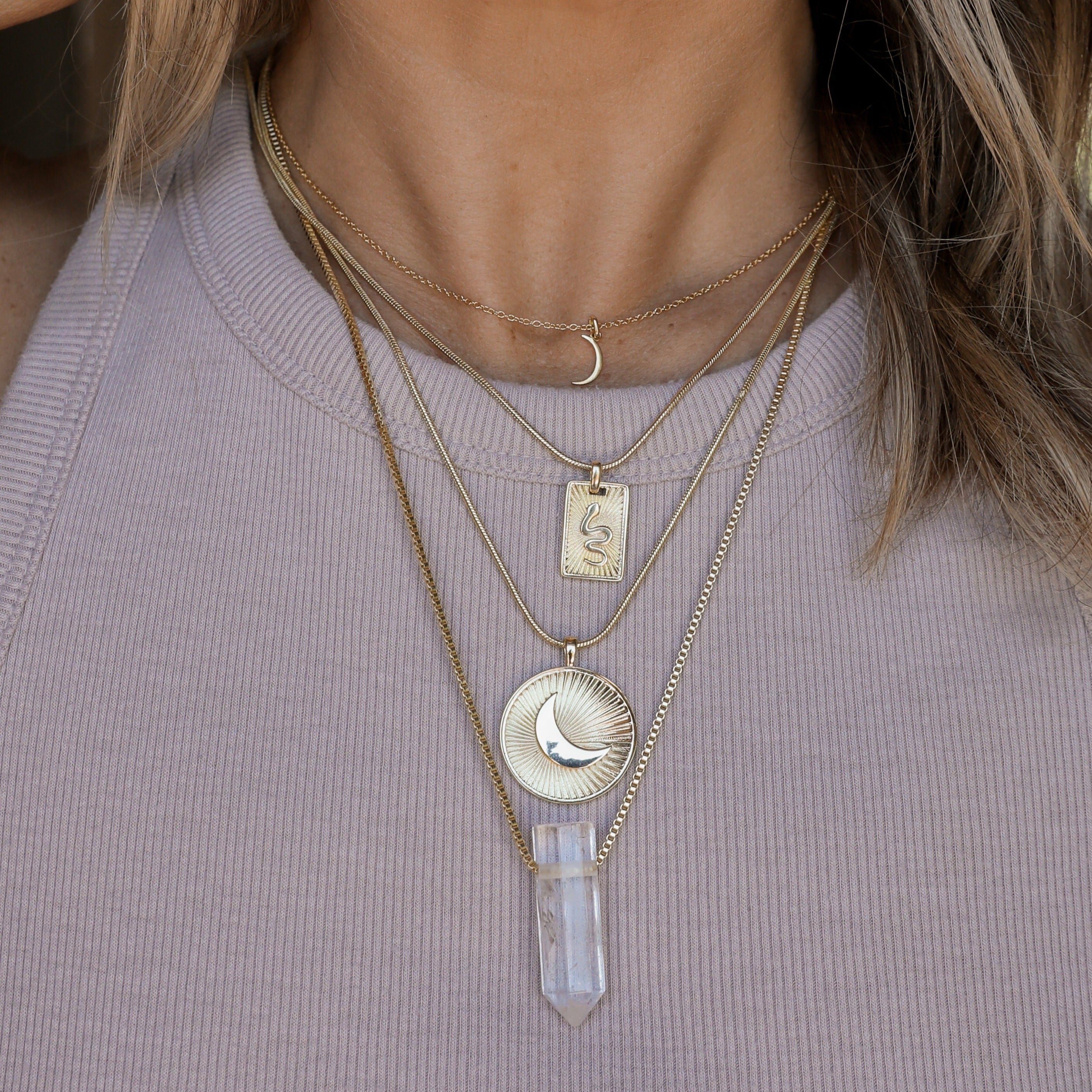 A close-up of a woman wearing a ribbed lavender tank top and four layered gold necklaces, including the Tiny Crescent Moon Necklace—an 18k gold plated piece with an adjustable chain, sun and snake, sun and moon charms, and a clear quartz crystal pendant.