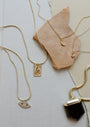 Four 18k gold plated necklaces—a Tiny Star Necklace, a crescent moon, a rectangular snake, and an eye with Cubic Zirconia charm—are displayed on textured neutral surfaces and tan stone.