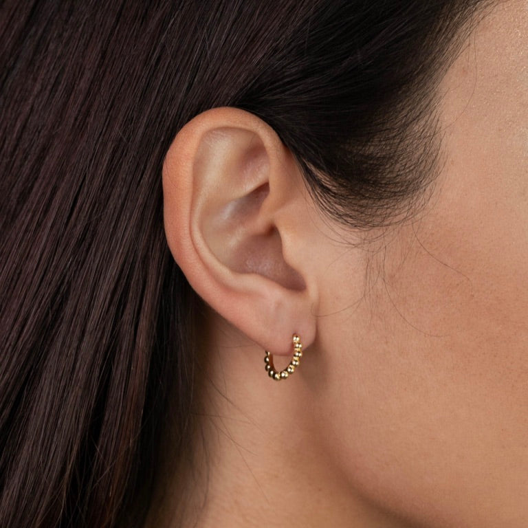 A close-up of a woman wearing the Small Ball Hoop: gold plated earrings featuring small ball details. Her straight dark hair is tucked back, highlighting the hoops. Sterling silver posts offer comfort and quality.