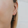 Close-up of a person’s ear wearing the Black Onyx Gemstone Drop - Focus earring, featuring modern gold plating and a sleek rectangular black onyx accent. A softly blurred background highlights its refined elegance.
