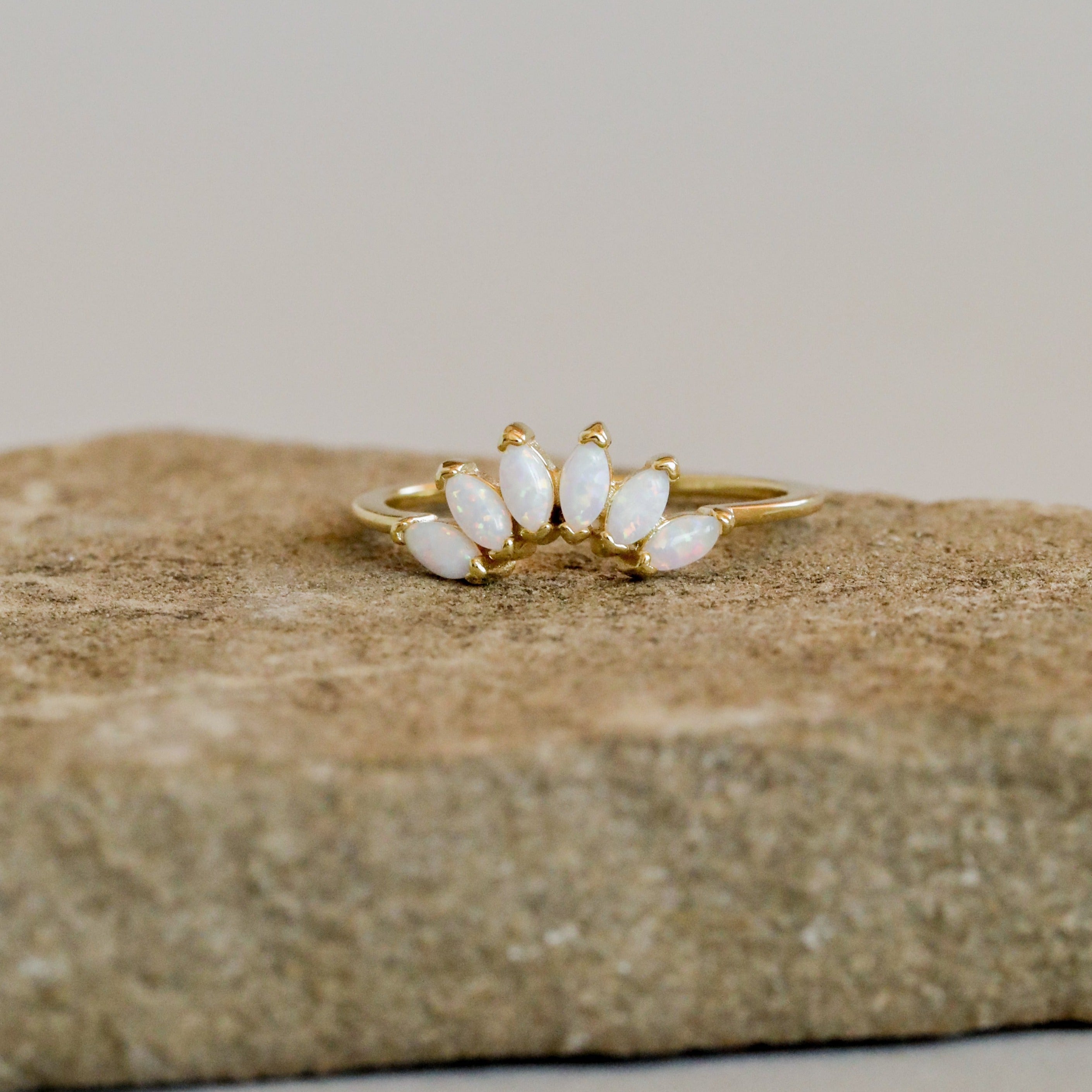 The Opal Crown Ring features five marquise-cut white opals in a graceful curve, set on an 18k gold plated band. Shown on a textured stone, it's an elegant piece ideal for stacking collections.