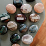 A selection of polished palm stones in various colors is displayed on a gray surface, including the Moonstone Palm Stone - Luck, each labeled and described with rectangular stickers.