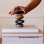 A hand balances the Moonstone Palm Stone—Luck, a smooth oval crystal, atop a stack of closed books in soft light, evoking a minimal and serene manifestation practice.