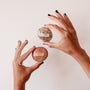 Two hands with black nail polish hold round Moonstone Palm Stones—one light brown, the other marbled white and beige. Each genuine crystal stone boosts your manifestation practice and is perfect for daily rituals. Product: Moonstone Palm Stone - Luck.