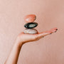 A hand with painted nails balances three smooth, stacked stones—including the Moonstone Palm Stone for Luck—against a soft pink background, evoking calm and manifestation.