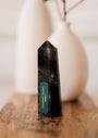 A Labradorite Crystal Point with a label stands on a wooden surface, radiating a protective aura. Two white ceramic vases appear blurred in the background.