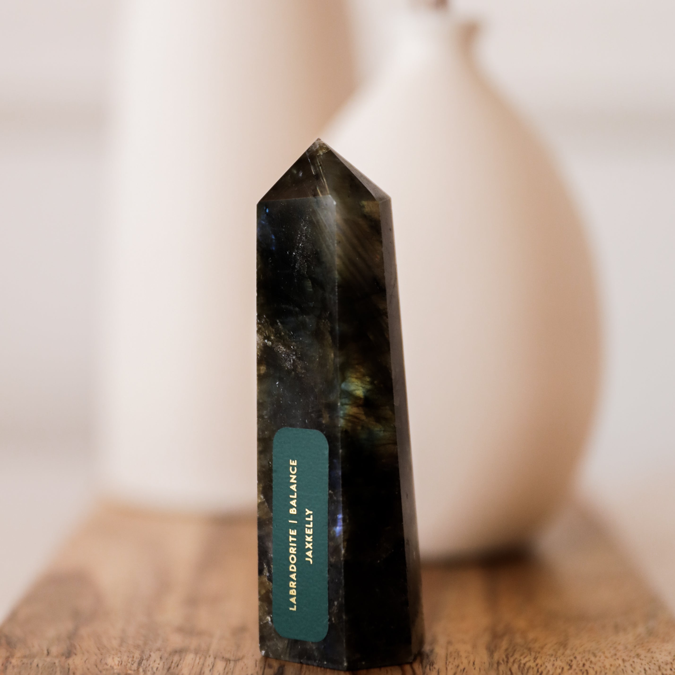 A Labradorite Crystal Point with a label stands on a wooden surface, radiating a protective aura. Two white ceramic vases appear blurred in the background.