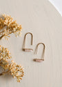 The Rose Quartz Gemstone Drop - Love earrings, featuring light pink stones and 18k gold plating, rest on a textured beige surface beside dried beige flowers.