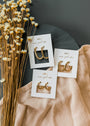 Three pairs of gold earrings, including the Small Rectangular Hoop with gold plating and sterling silver posts, are displayed on white cards over beige fabric with dried flowers against a dark background.