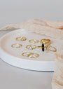 A white round tray displays eight Simple Stud Rings, each 18k gold plated with assorted designs—featuring gemstones, pearls, and unique shapes—while beige textured fabric is partially draped around the tray.