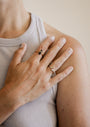 A close-up of a hand resting on a shoulder, wearing a sleeveless ribbed light gray top and two rings—one is the Simple Stud Ring in sleek 18k gold plate, the other is black. The skin is light-toned and nails are natural.