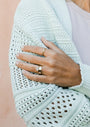 A person crosses their arms in a light sweater, displaying manicured nails and three gold rings—including the delicate Lambda Ring—on their fingers. The focus is on their hands and rings.