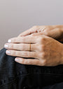 A close-up of a person’s hand resting on their knee, wearing the Half Pavé Ring—a minimalist gold plated ring—on the ring finger. Their nails are neatly manicured with light polish, and they’re dressed in dark clothing.
