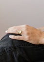 A hand with light skin and short, neatly manicured nails rests on a bent knee, adorned with the Mother of Pearl Signet Ring—a gold ring featuring a round white mother of pearl stone—paired with dark pants.