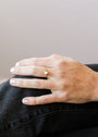 A hand with light nail polish rests on dark jeans, showcasing an elegant Opal Burst Ring—an 18k gold plated piece featuring a single diamond on the ring finger.