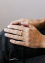 A close-up of hands resting on a leg, showcasing neatly manicured nails and the Ridged Ring—an elegant stackable band with 18k gold plating—on the ring finger. The softly blurred background features neutral tones.
