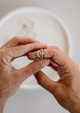 A person holds a stack of Simple Stud Rings, 18k gold plated with unique designs—flower, arrow, and shimmering cubic zirconia—against a softly blurred background with a white circular dish.