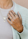 A person in a light knit sweater and beige top displays gold necklaces and stacking rings, including the Opal Crown Ring—an 18k gold plated ring with a black stone—resting their hand near their collarbone.