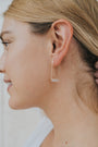 Close-up of a woman's ear wearing the Rose Quartz Gemstone Drop - Love earring, featuring 18k gold plating and a rose quartz rectangular stone. She has light skin and blonde hair; the background is softly blurred.