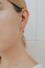 Close-up of a person’s ear wearing the Rose Quartz Gemstone Drop - Love, a minimalist drop earring with 18k gold plating and a small rose quartz bead. The background is neutral, with the wearer’s light skin and blonde hair softly visible.