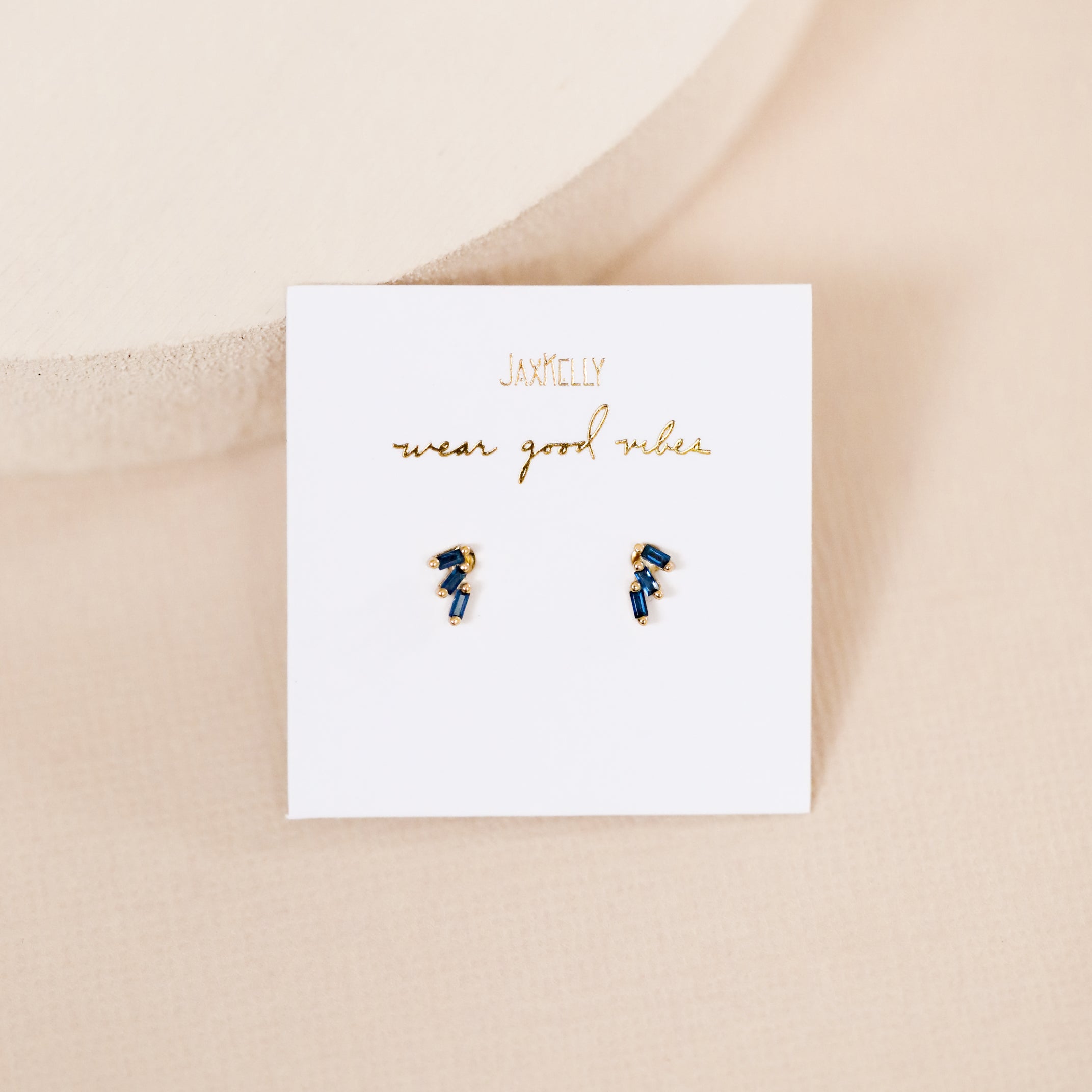 The Offset Trio - Sapphire earrings are displayed on a white card with gold "JaxKelly wear good vibes" lettering, set against a soft beige background.