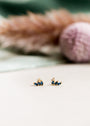 The Offset Trio - Sapphire features small gold plated stud earrings with blue sapphire CZ stones, displayed on a light surface accented by soft-focus neutral and pink decor in the background.